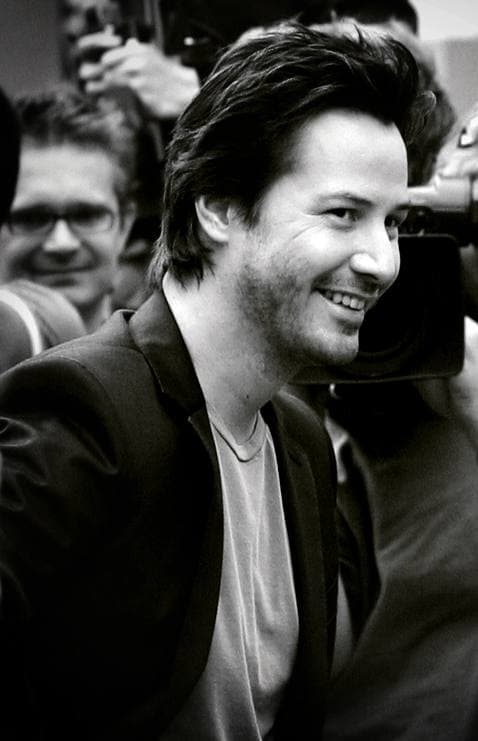 Keanu Reeves smiling, dark hair swept back, in a black-and-white photograph