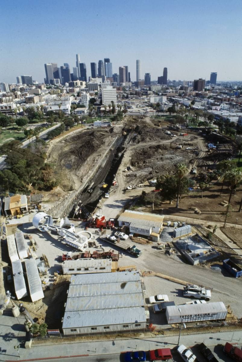 An aerial view of a large excavation with cranes, the downtown skyline behind