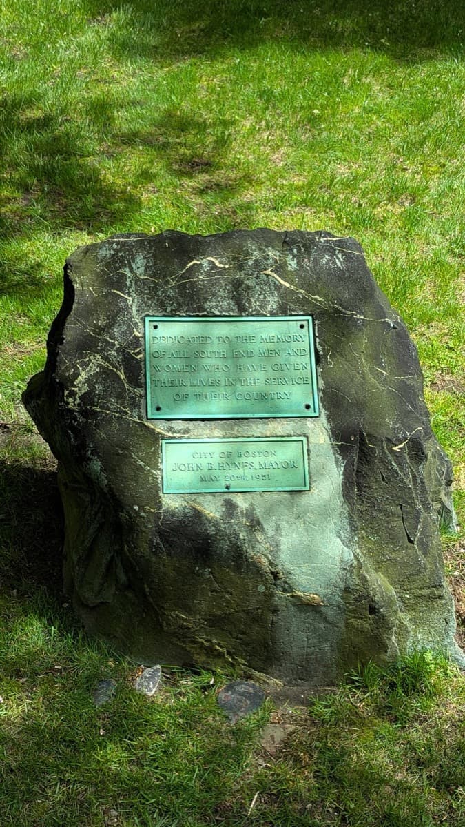 A small stone memorial standing in a garden with trees and brick houses behind
