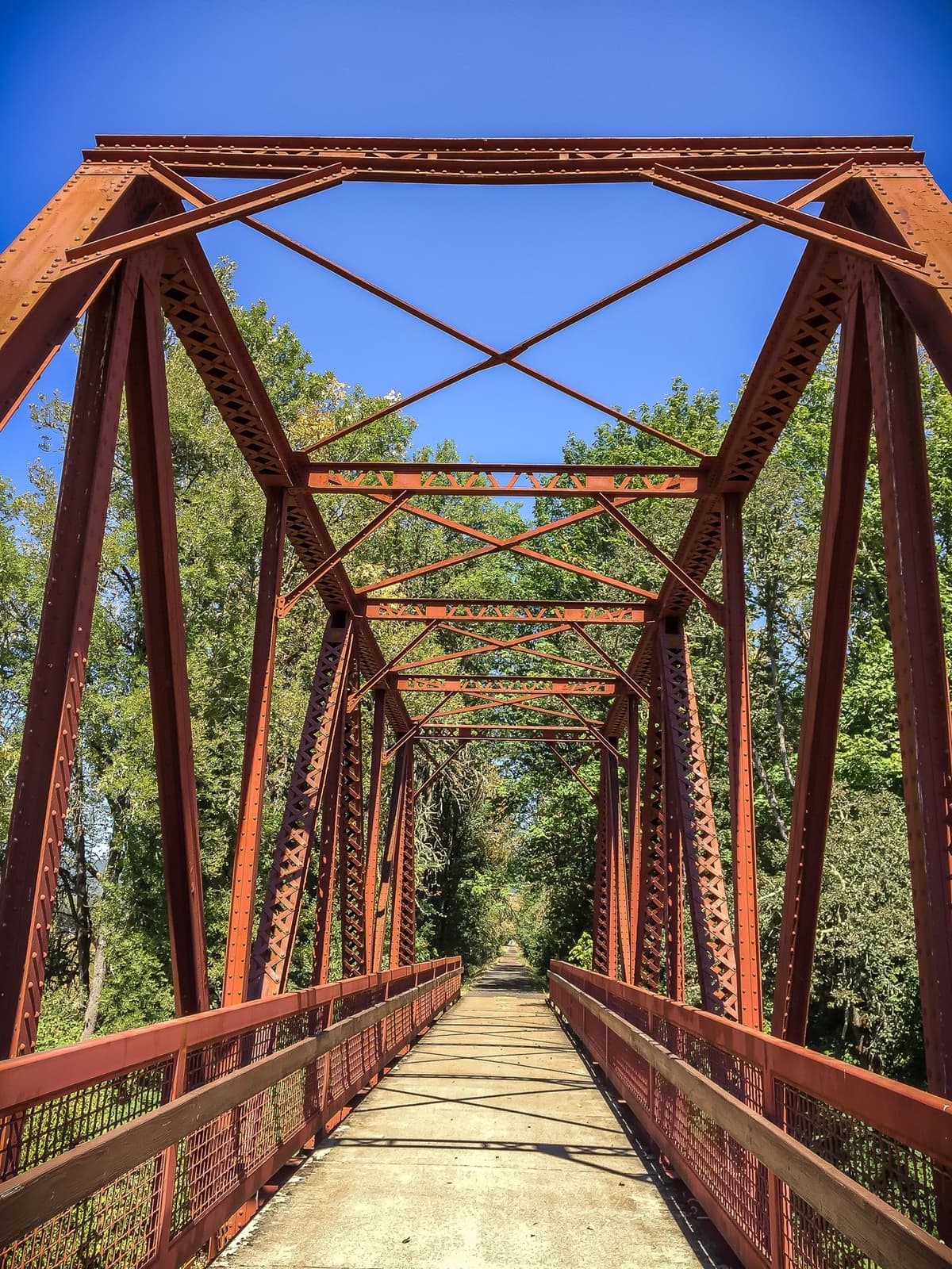 Looking through a rust-red steel truss bridge on a paved trail, trees on both sides, blue sky