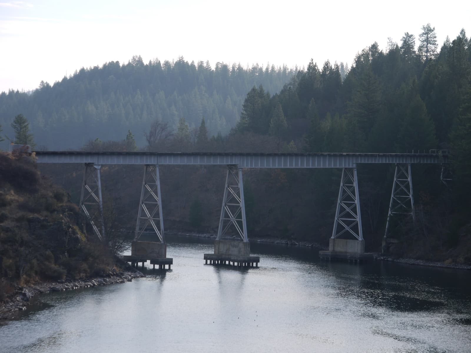 A tall white steel trestle on concrete piers crossing a still, dark lake between pine-covered hills