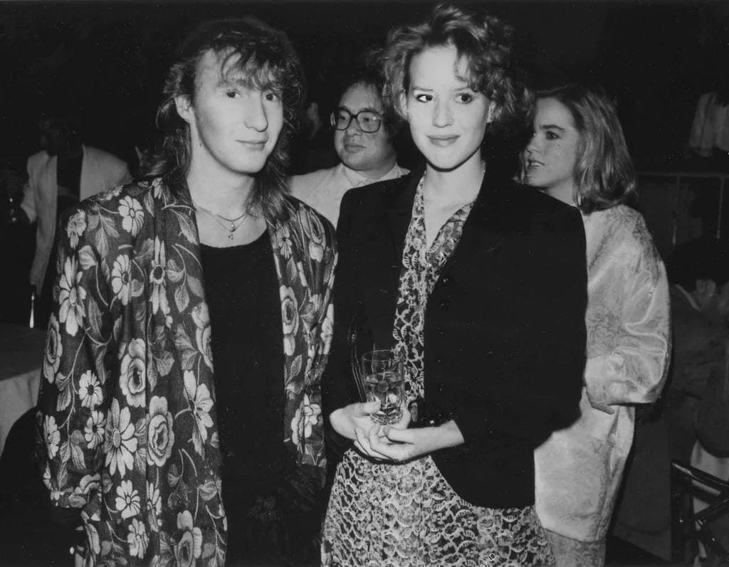 Molly Ringwald in a dark jacket beside Julian Lennon in a floral jacket at a crowded party, black and white