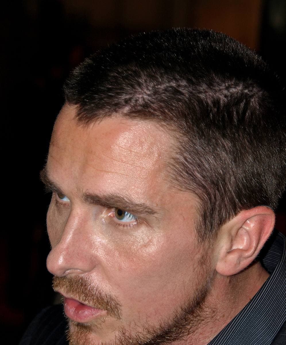 Christian Bale with cropped hair and stubble looking to one side under warm light