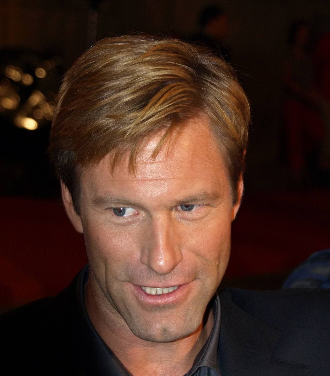 Aaron Eckhart smiling, swept-back fair hair, dark shirt