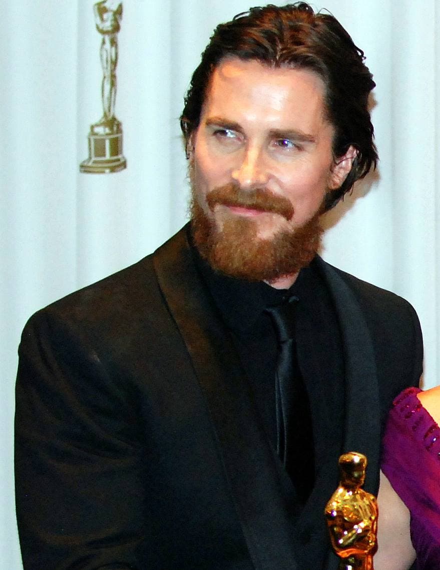 Christian Bale with long hair and a full beard holding an Oscar