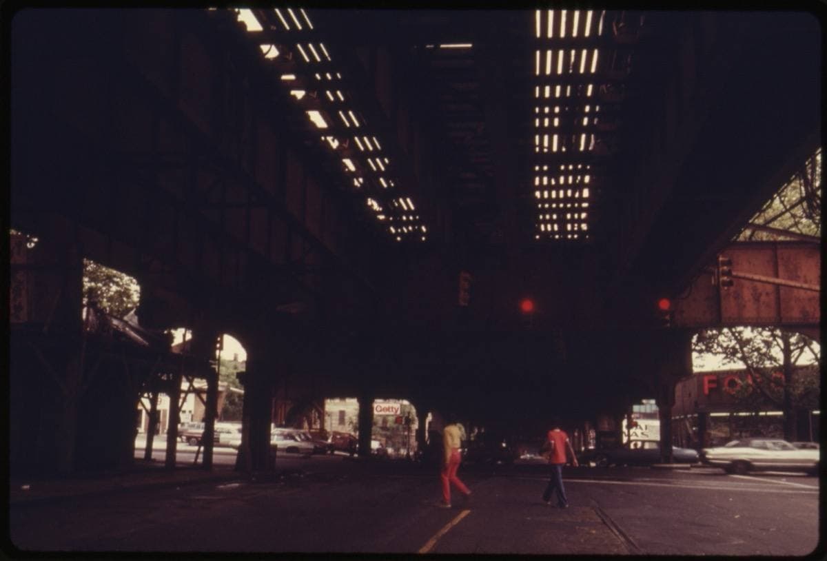 Colour photograph from 1974 of the dark steel underside of an elevated train line over a Brooklyn street