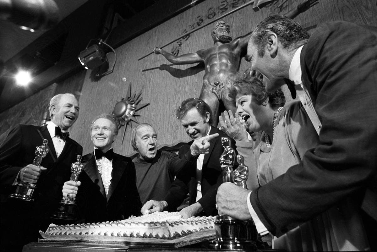 Black-and-white photograph of a group of men in suits holding Oscar statuettes around a cake
