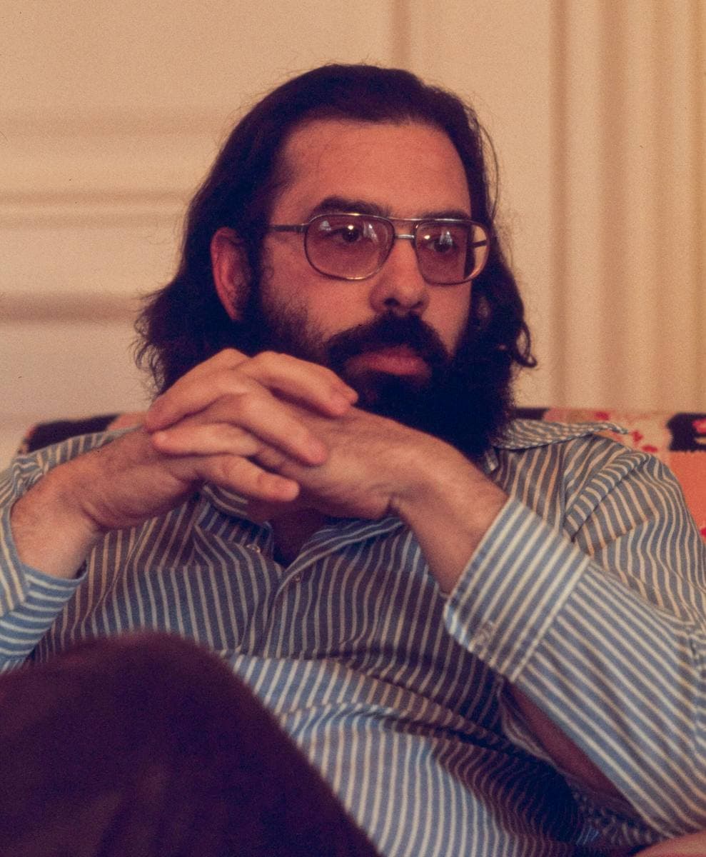 A bearded man with long dark hair and glasses, hands clasped, in a striped shirt