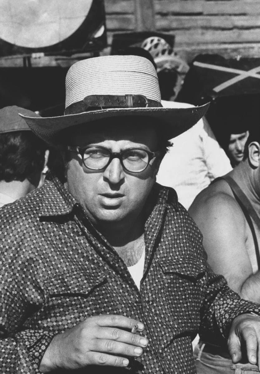 Sergio Leone in glasses and a straw hat with a cigarette, crew behind him