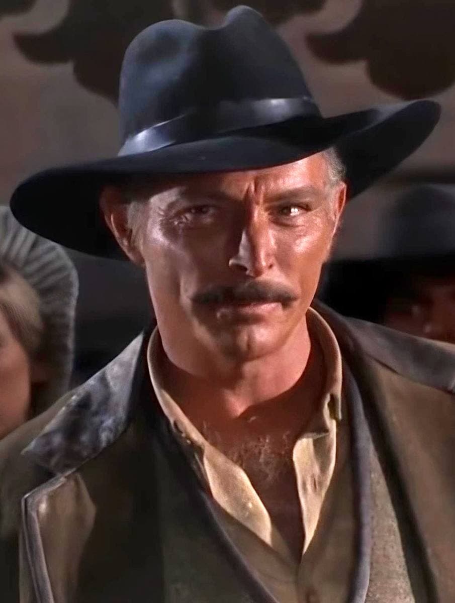 Lee Van Cleef in a black hat and brown coat, staring hard at the camera, moustache and narrowed eyes