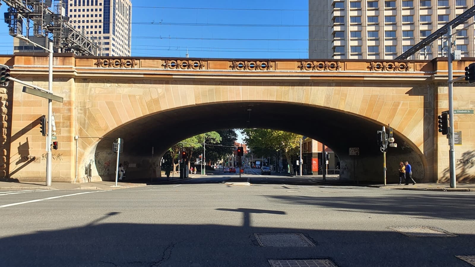 A sandstone railway bridge with a wide arch over an empty street in sunshine
