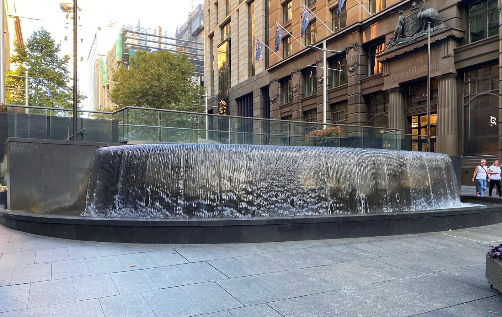 A wide, low round fountain with water spilling over its rim, in a paved square lined with sandstone buildings