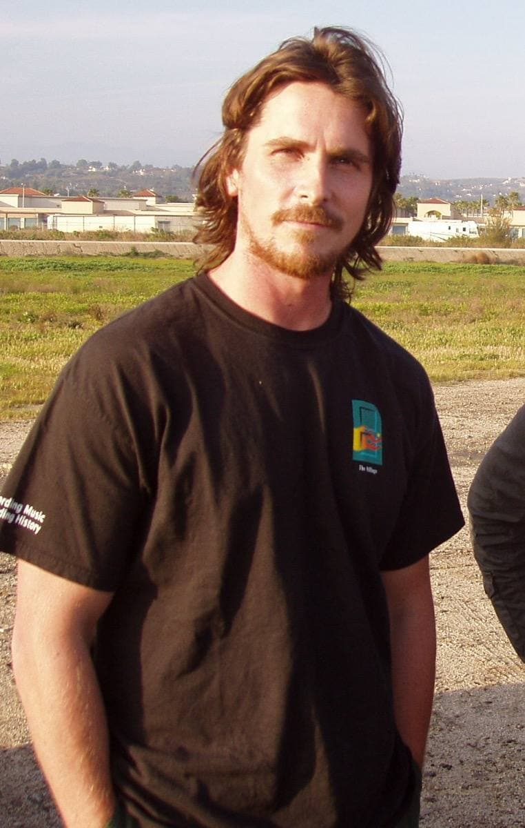 Christian Bale with long hair and a beard, in a brown T-shirt, outdoors