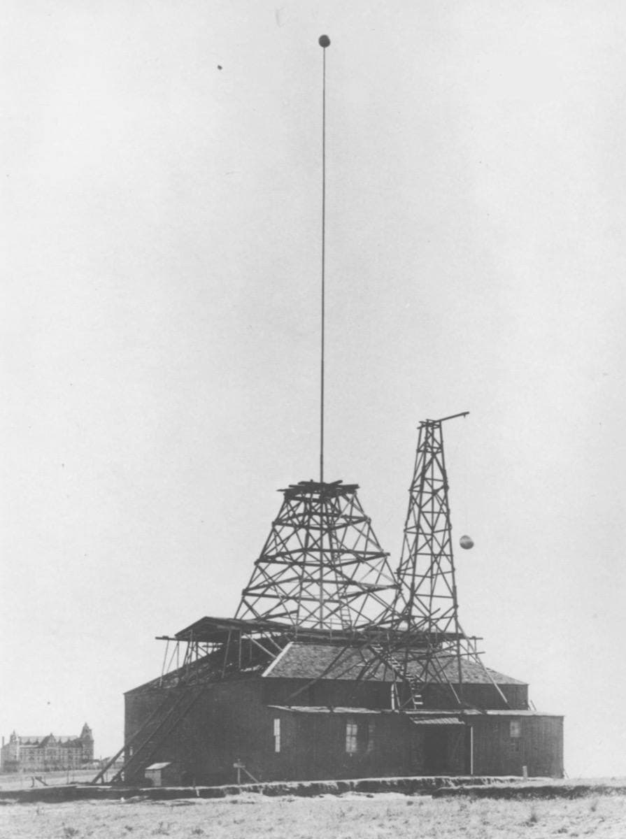 A timber shed with a lattice tower and a tall mast on bare ground
