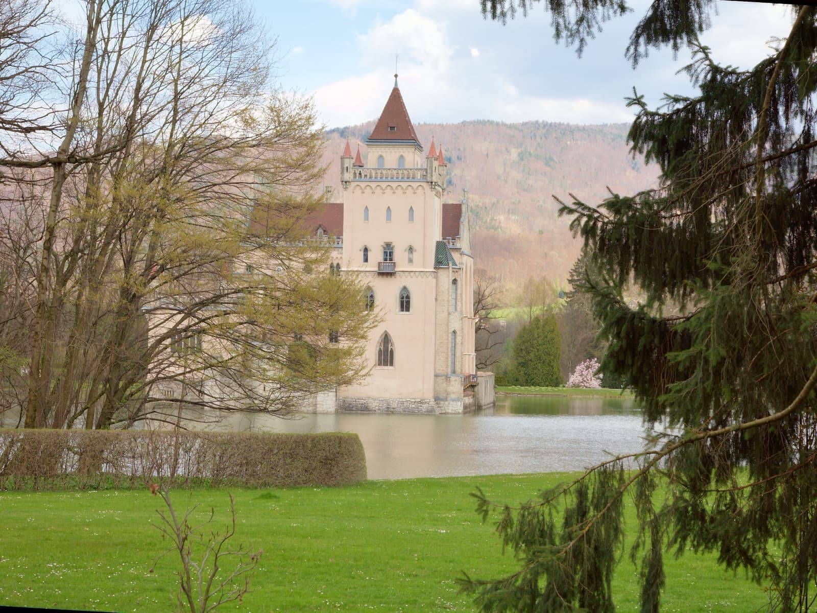 A pale gothic tower standing in a pond, behind trees