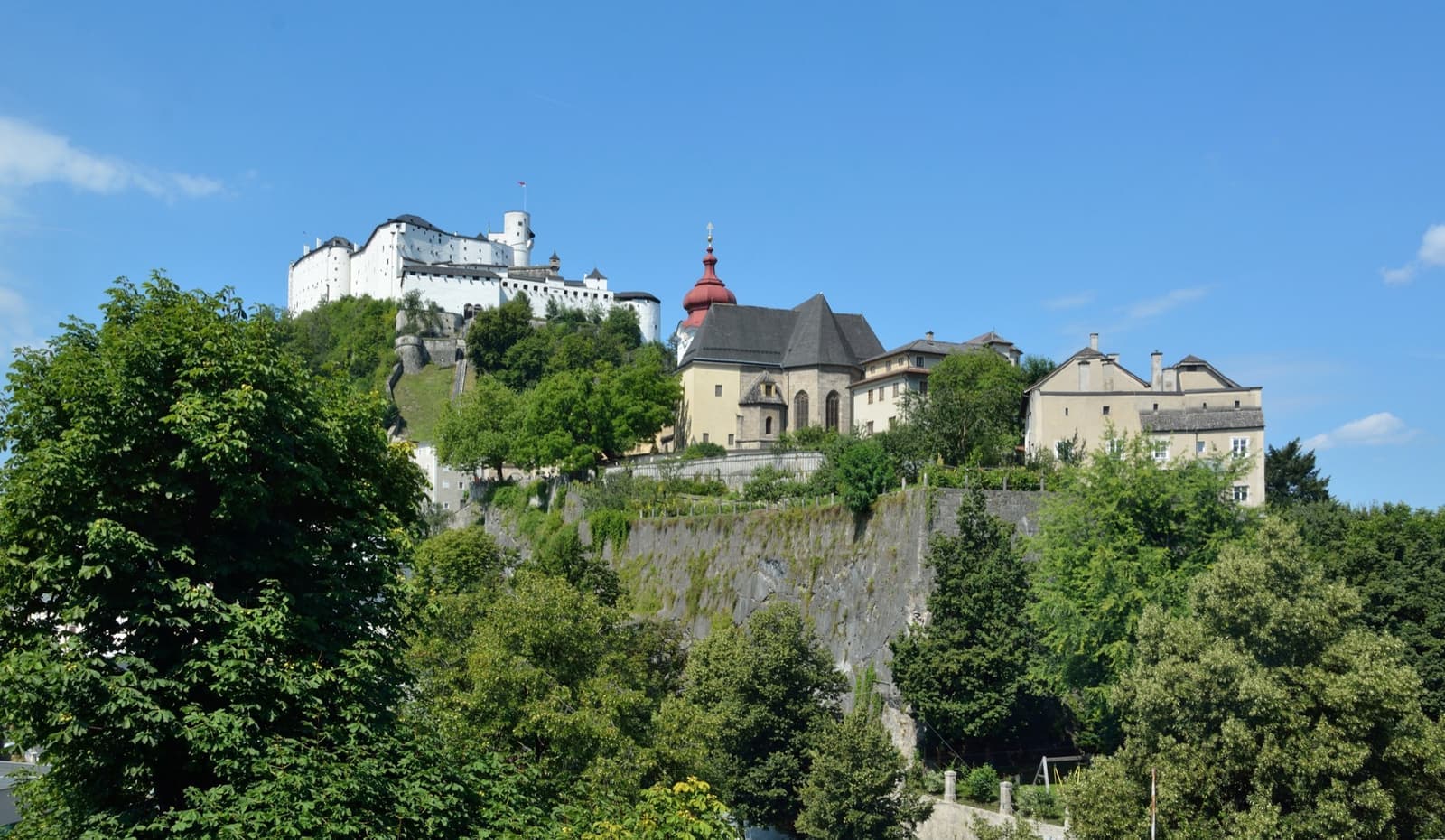 A convent with a red onion-domed tower on a wooded cliff, a fortress above