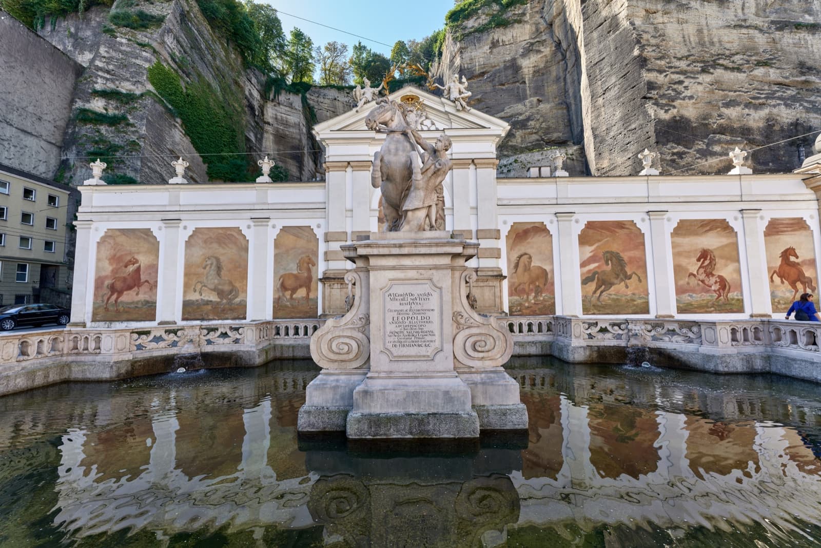 A baroque horse trough with painted horses on the wall behind a statue