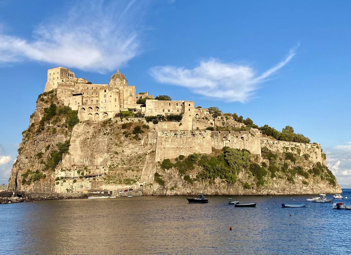 A castle on a rocky islet joined to the shore by a long stone causeway, over a blue sea