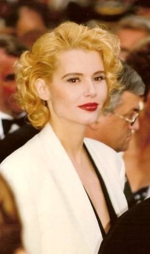 Geena Davis with curled blonde hair, red lipstick and a white jacket among a crowd