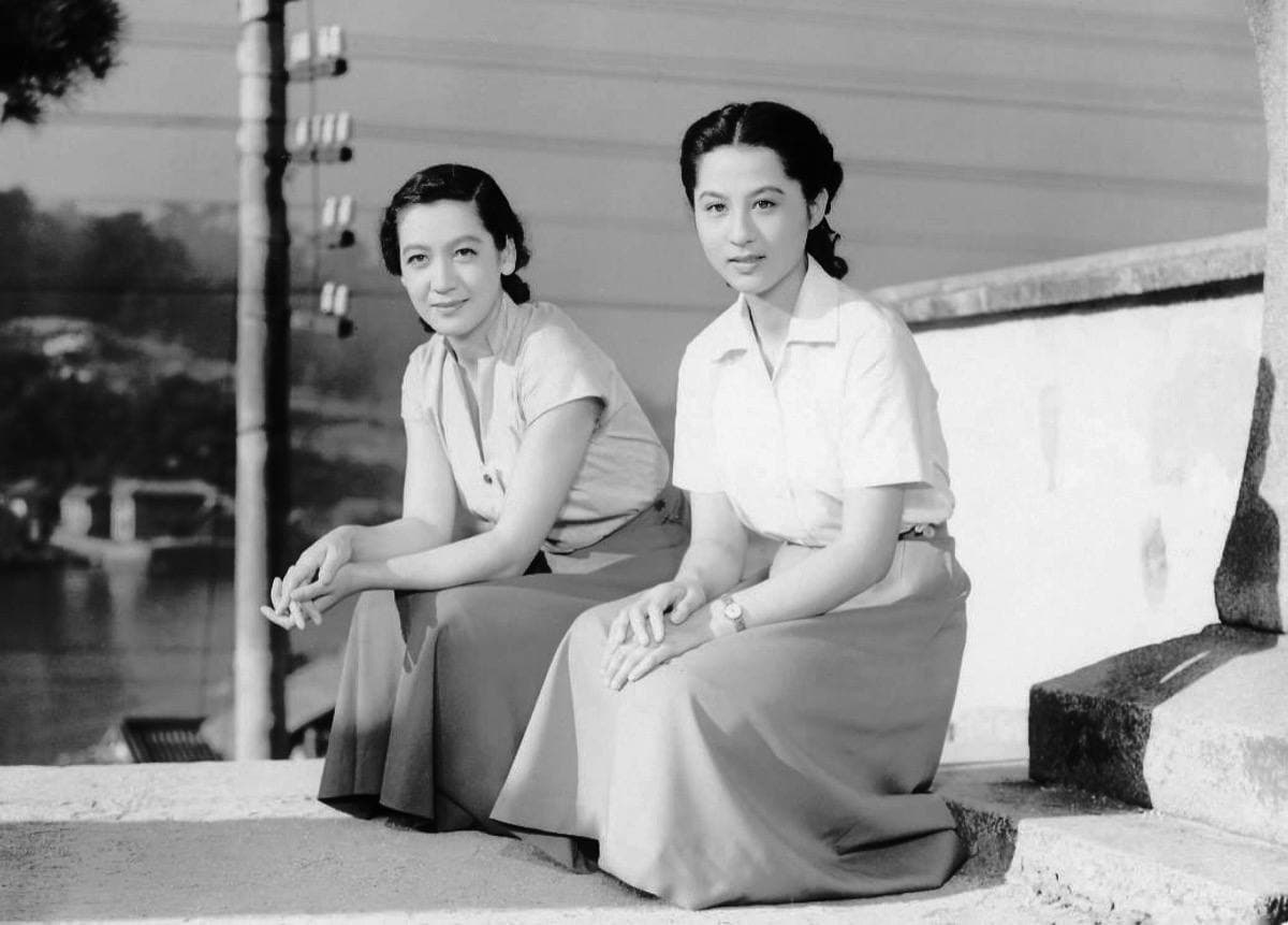 Two young women sit side by side on a stone step above the water, smiling