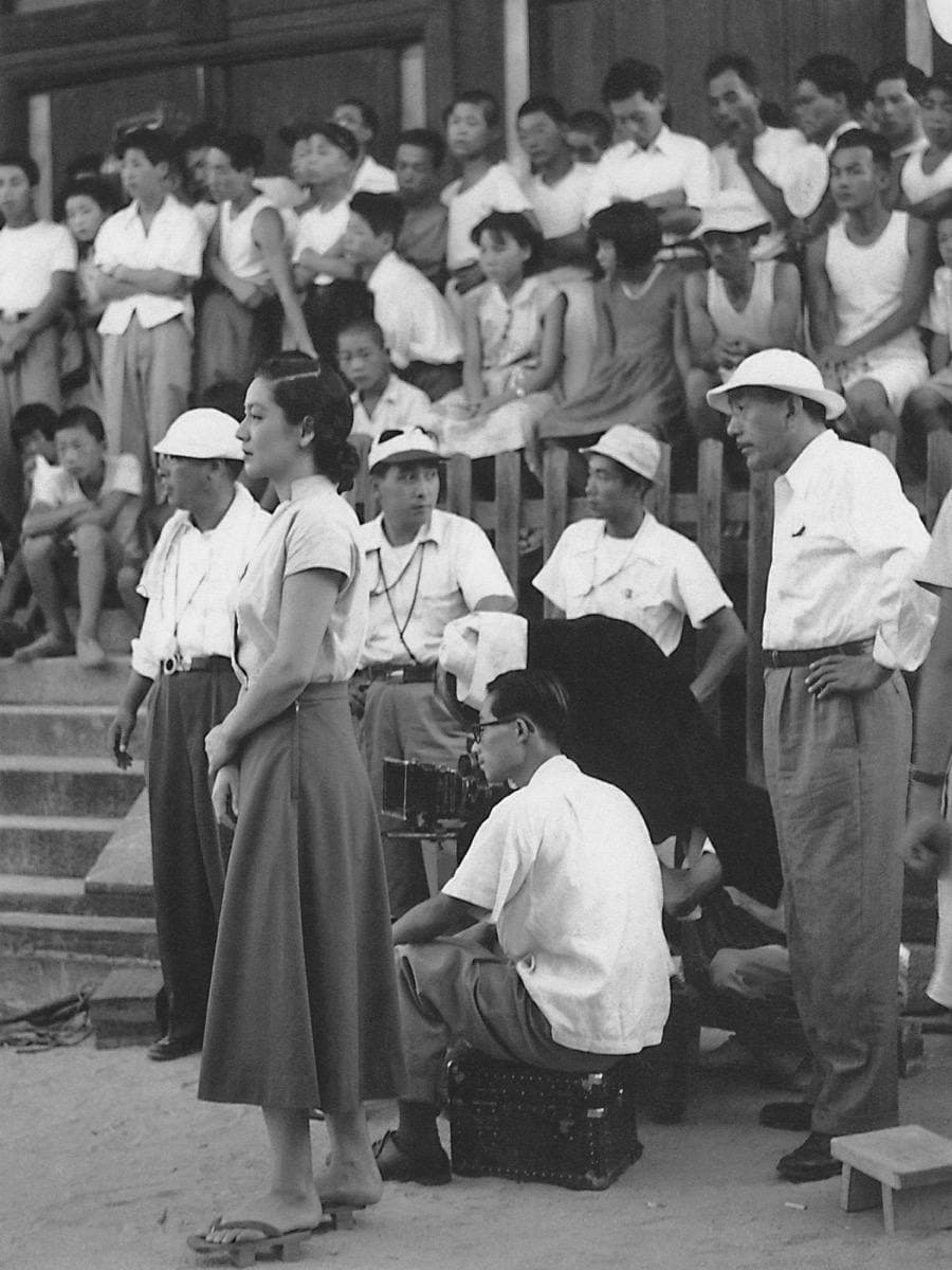 A film crew in white shirts and hats on temple steps, a crowd of onlookers behind them