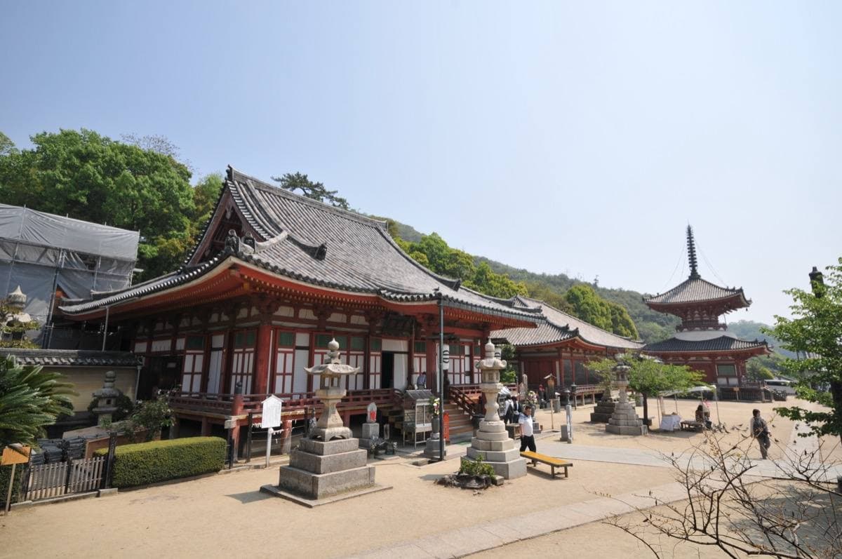A red-pillared temple hall and a two-storey pagoda on a sunny gravel precinct