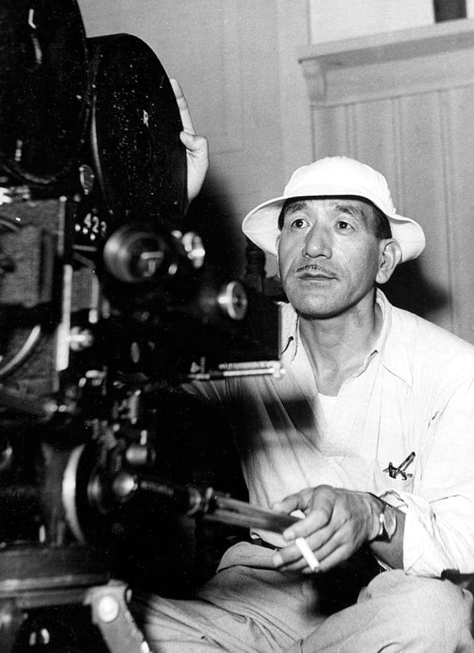 Yasujirō Ozu in a white sun hat, sitting low beside a film camera