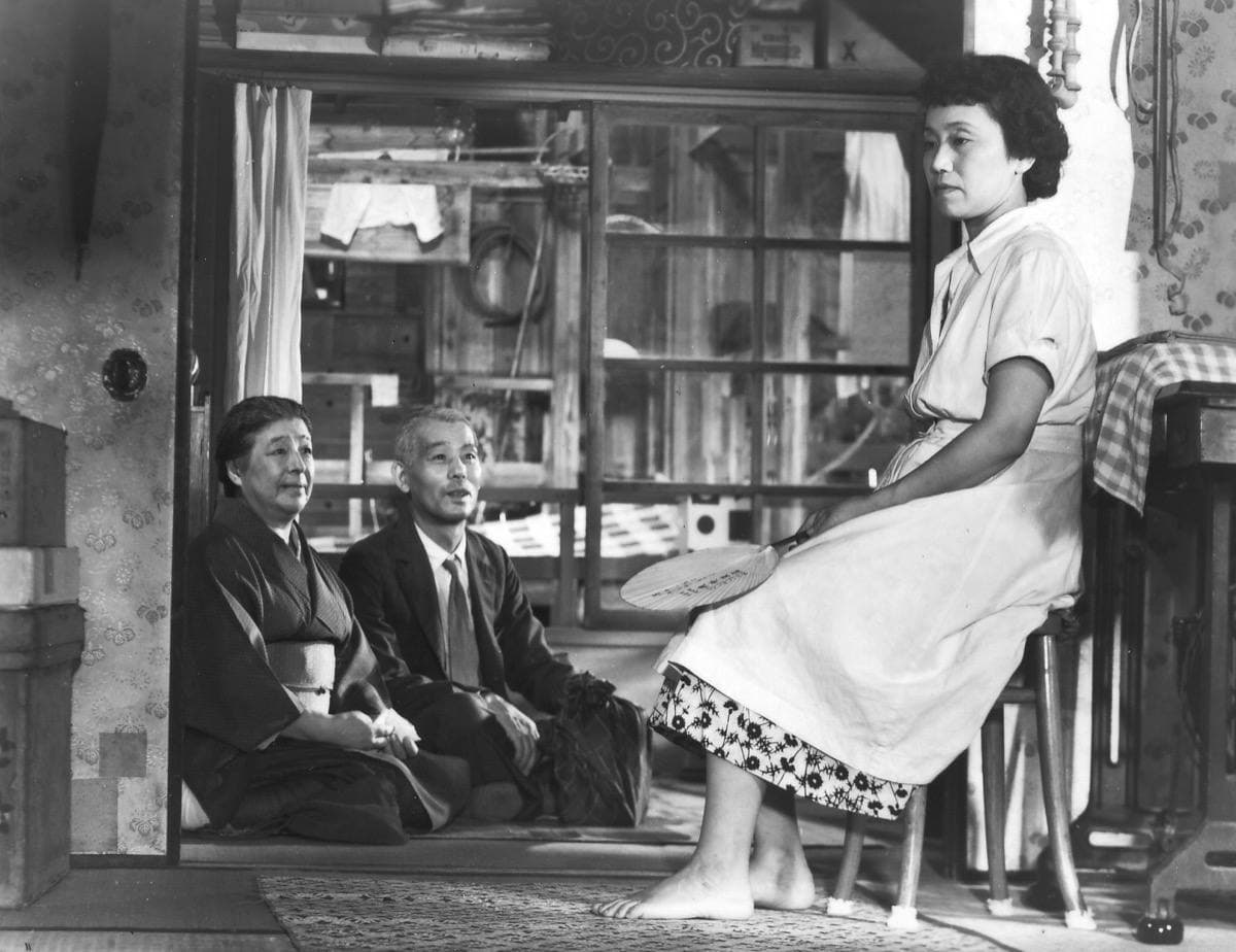 An old couple sit on the floor of a shop while a woman in a white dress sits on a chair beside them