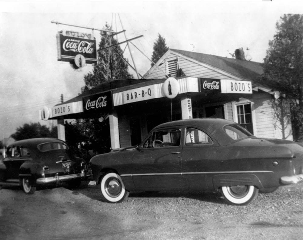 A black-and-white photograph of a small roadside barbecue stand with hand-painted signs