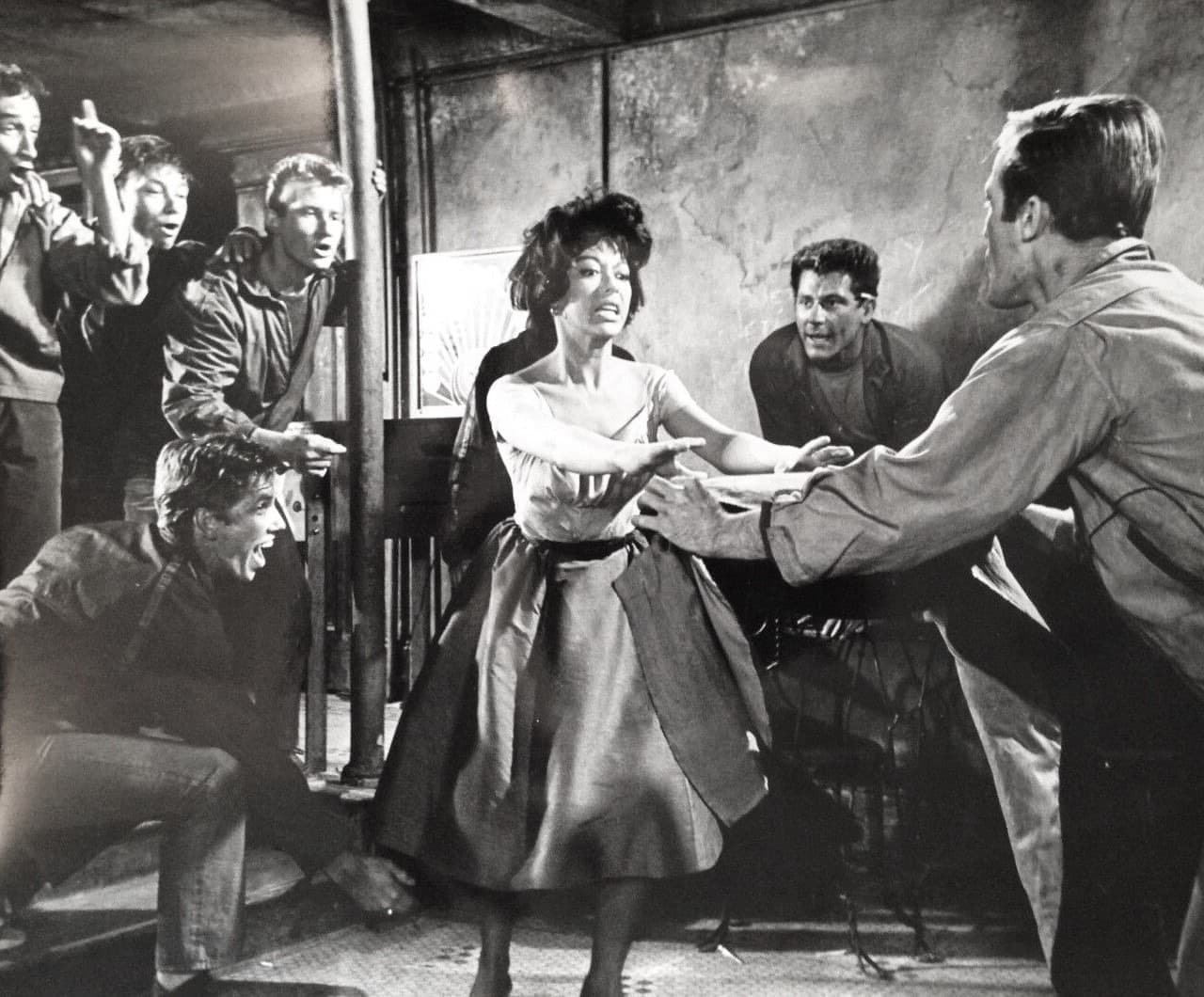 Rita Moreno in a dark dress pushed between young men in a cramped room