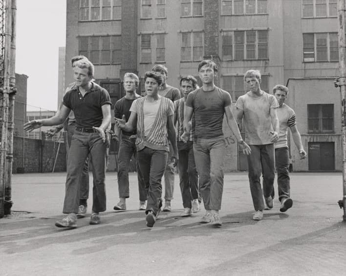 A line of young men in jeans and T-shirts walking towards the camera down a city street, tenements behind