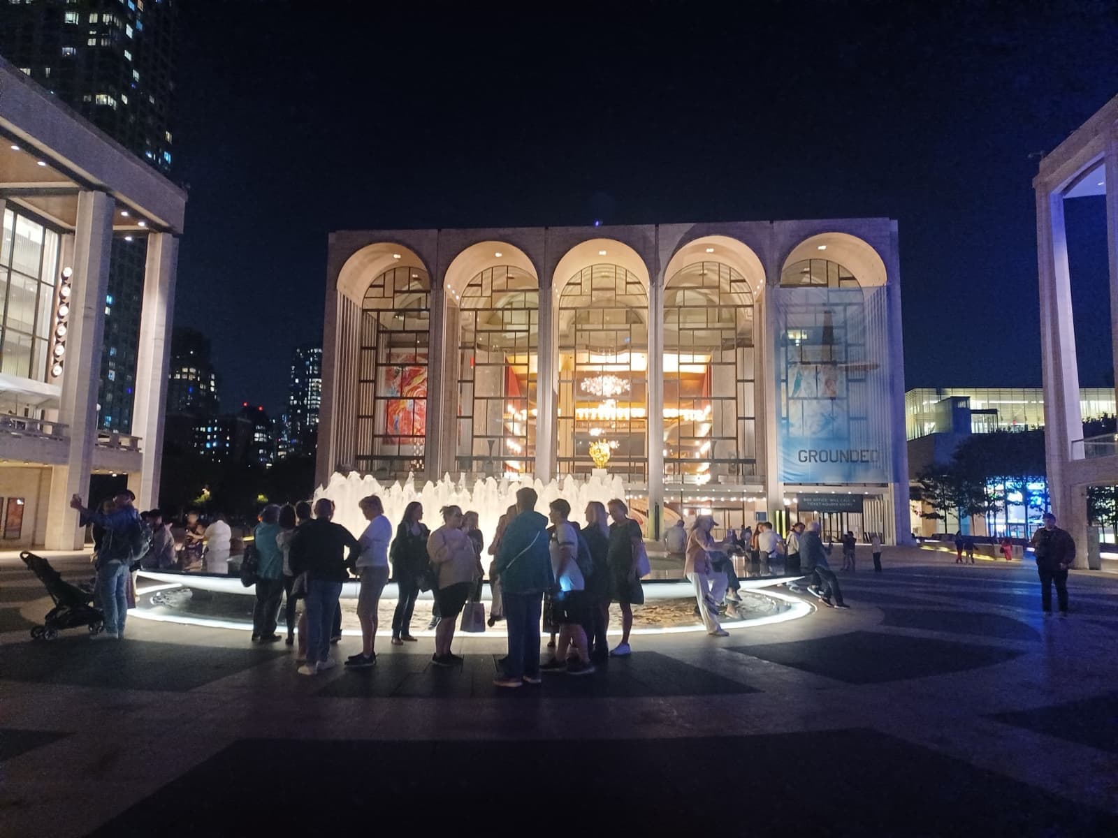 The arched glass front of the Metropolitan Opera House lit at night behind the plaza fountain