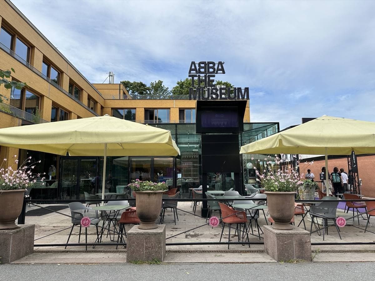 A low ochre building with tall black lettering over the entrance, café umbrellas in front