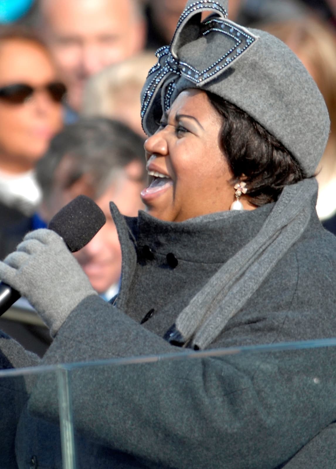 Aretha Franklin in a grey coat and a grey hat with a large bow, singing at a microphone outdoors