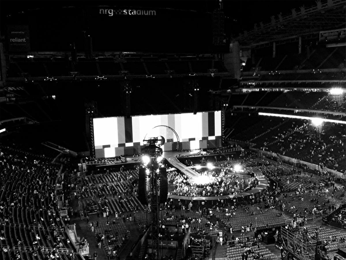 A black and white view across a packed stadium towards a lit stage and screen