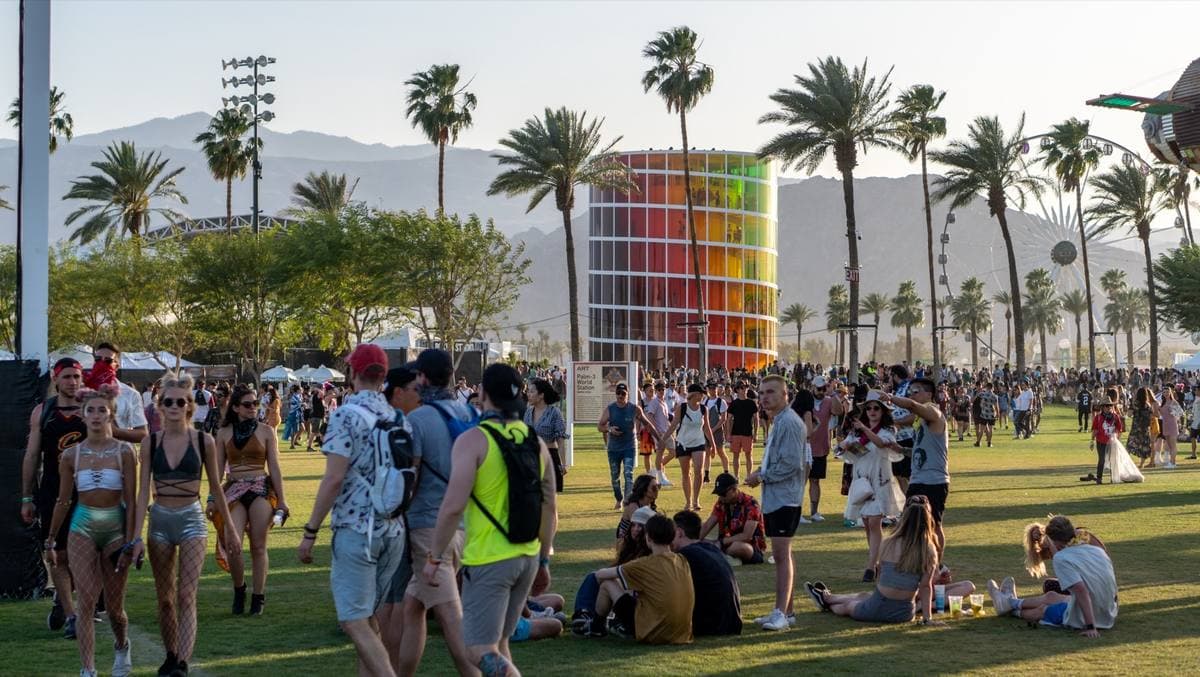 Festival-goers on a green lawn among palm trees, a rainbow-coloured tower behind them and mountains beyond