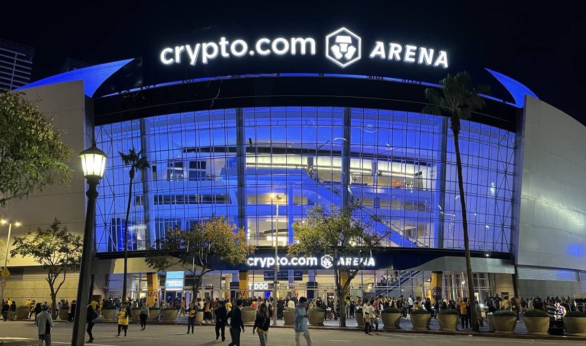 An arena with a glass front lit blue at night, people walking past palm trees outside