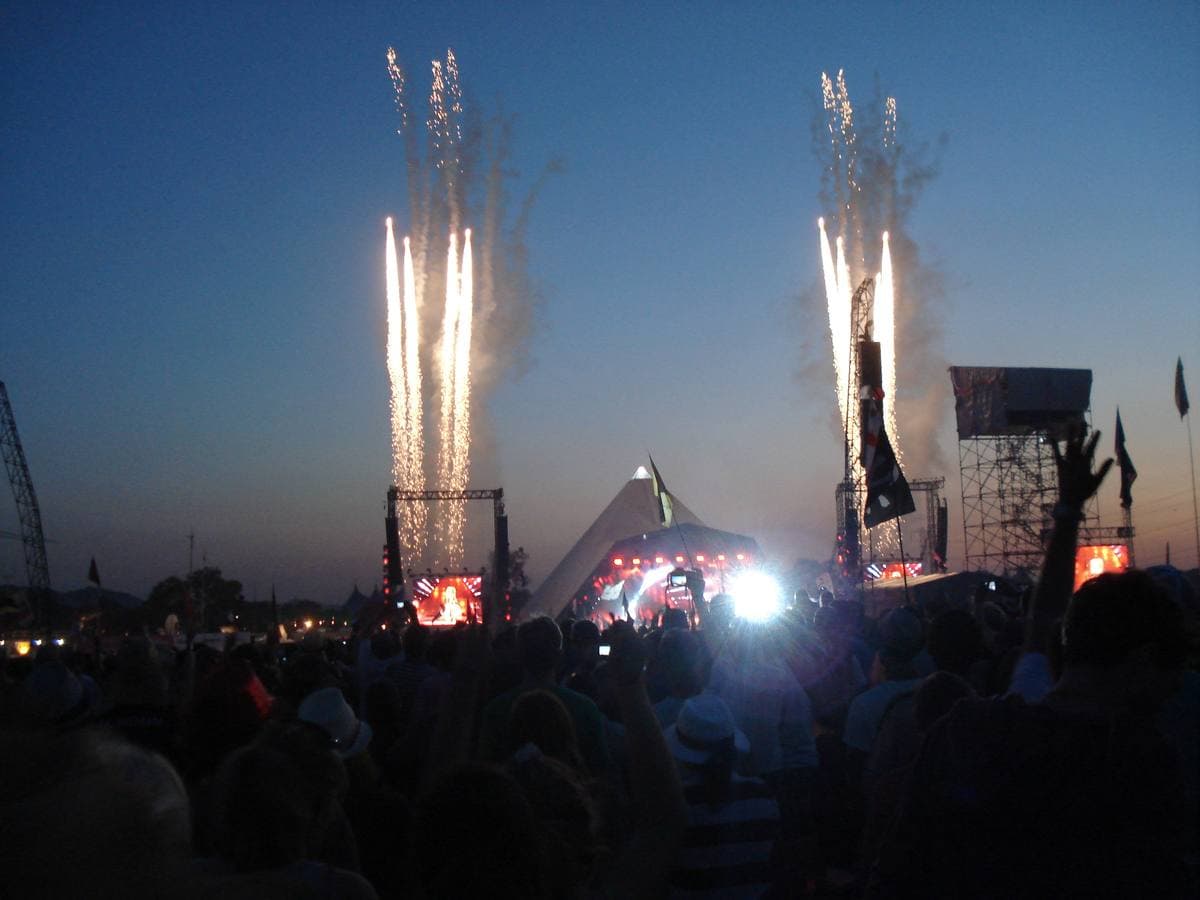 Fireworks shoot up either side of a stage at dusk above a dark crowd