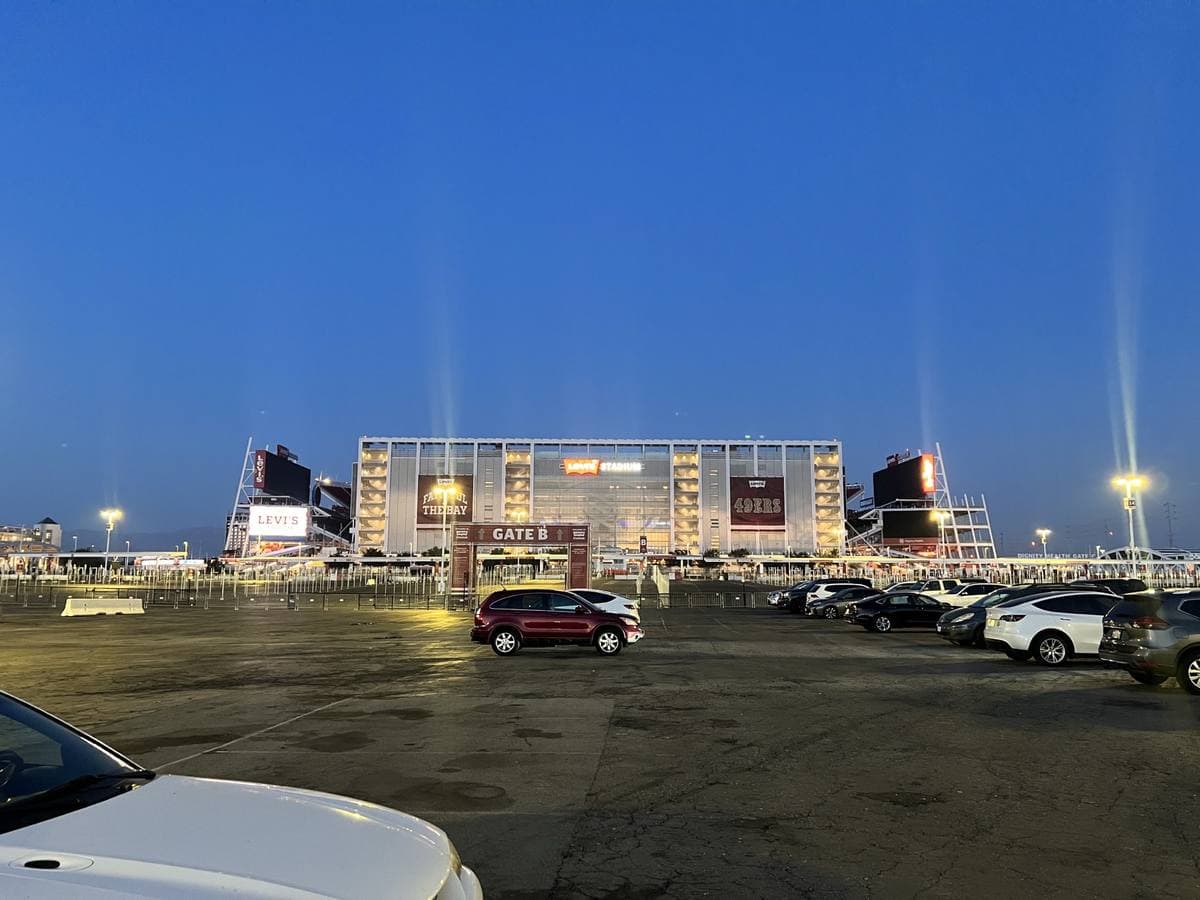 A stadium entrance lit up at dusk across a car park, searchlights in the sky