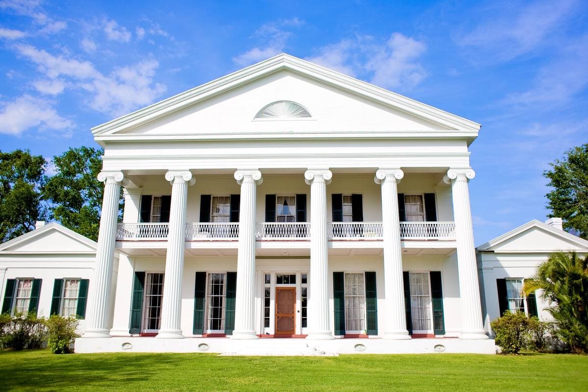 A white Greek Revival house with six tall columns and green shutters on a lawn