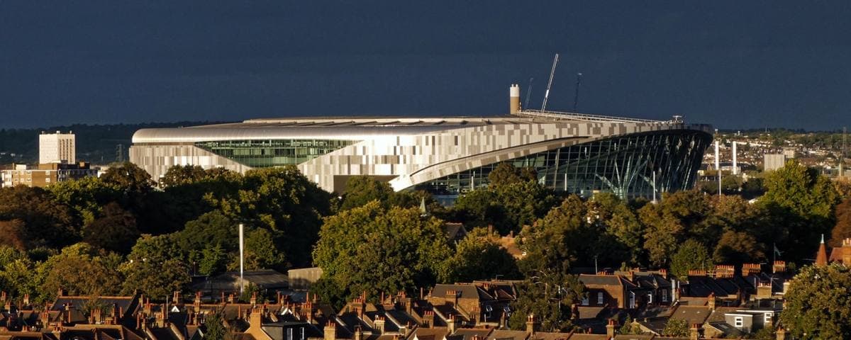 A large modern stadium catching low evening sun above the trees and rooftops of north London