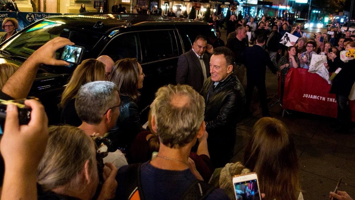 A crowd of people holding up phones around Bruce Springsteen at night on a Manhattan pavement
