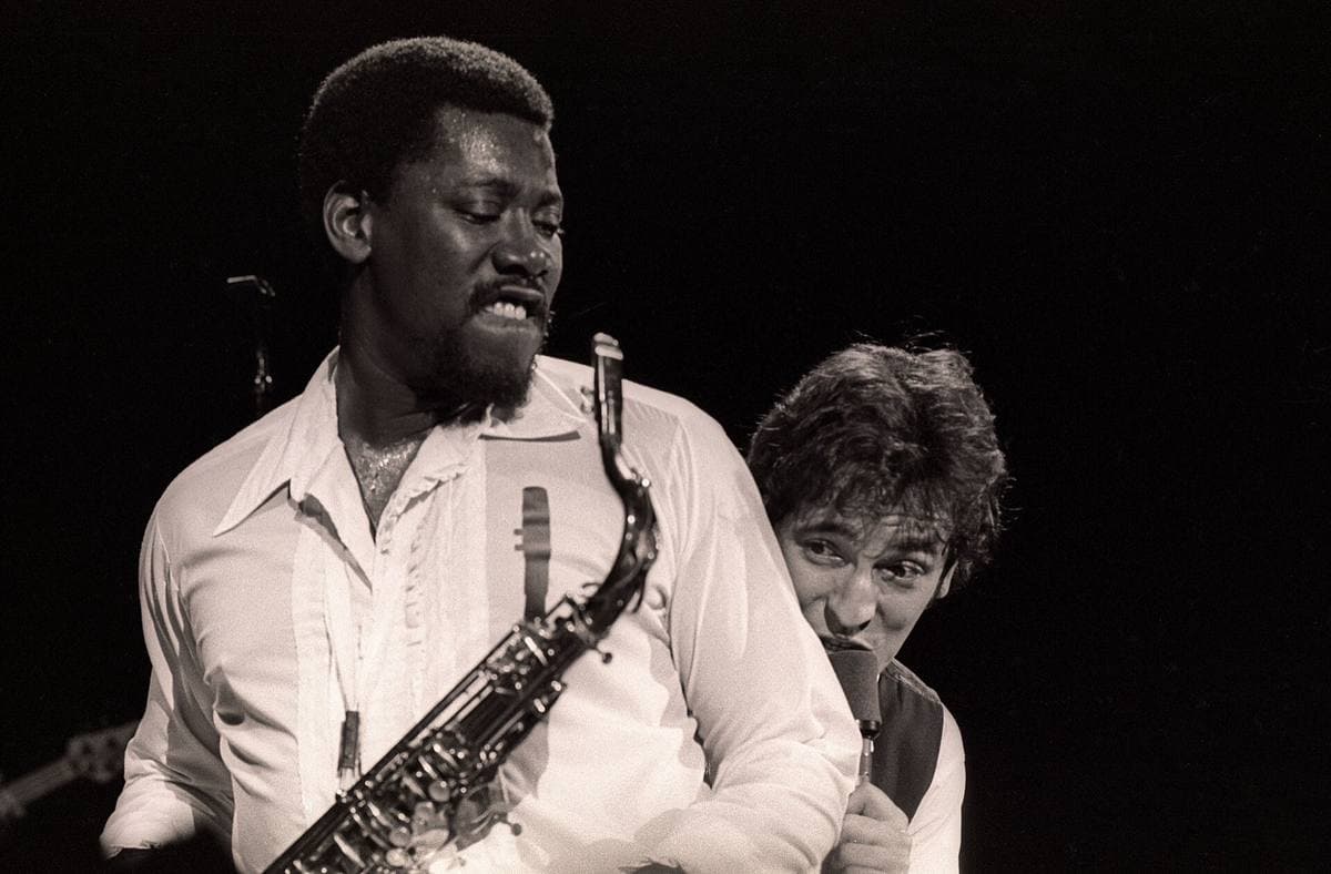 Clarence Clemons in a white shirt playing saxophone while Bruce Springsteen leans in grinning behind his shoulder, black and white