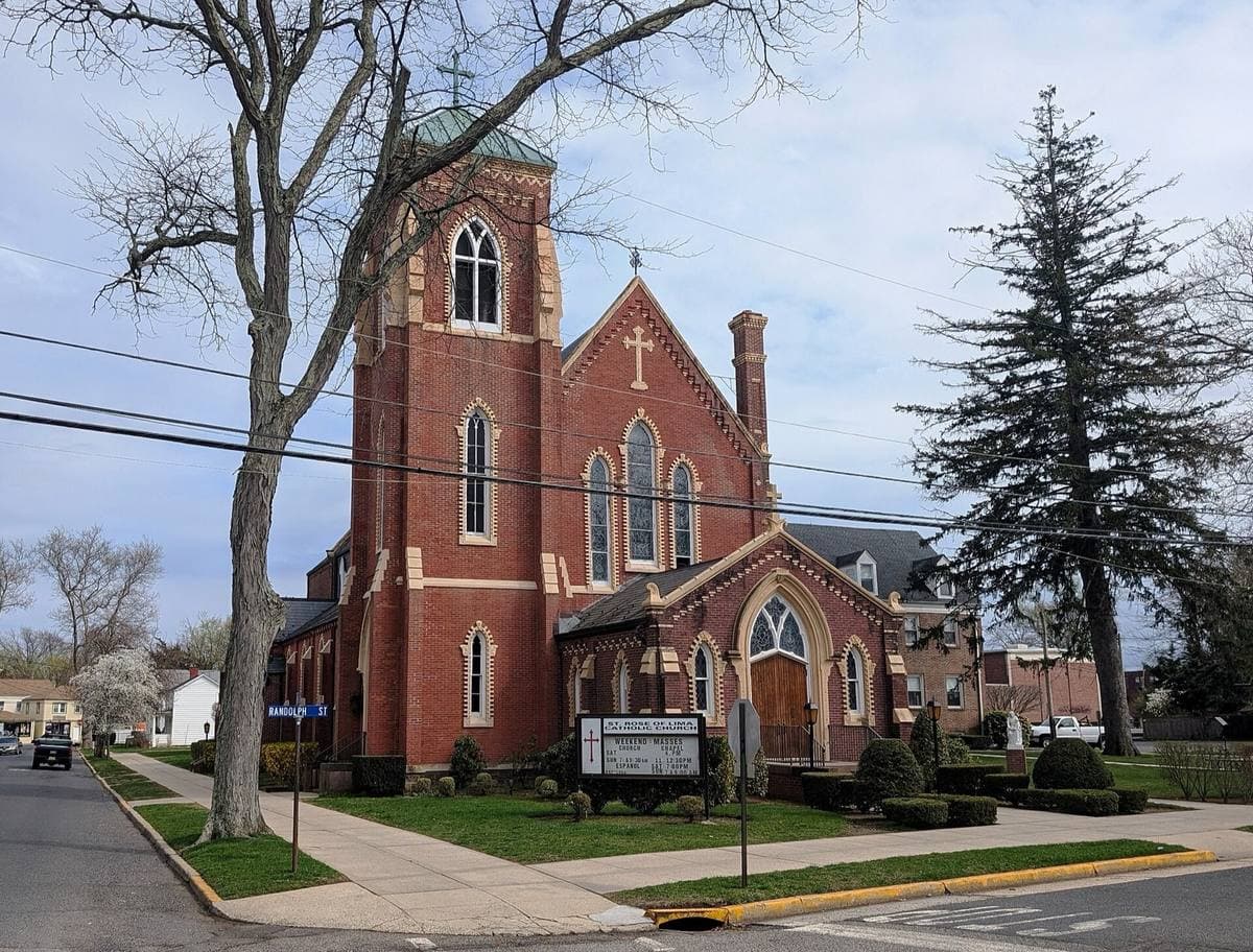 A red-brick Gothic church with a tall spire behind a lawn and a tree on a small-town street