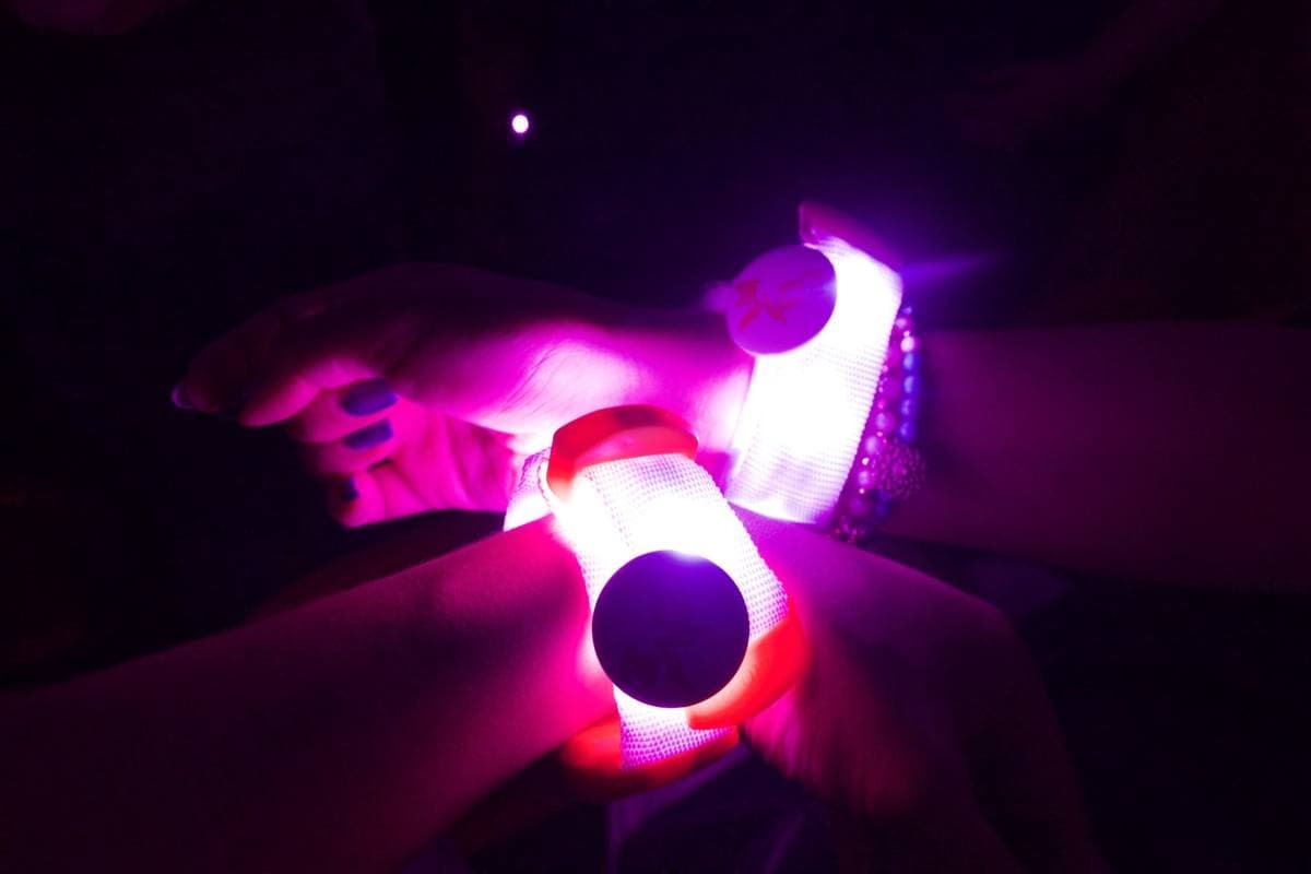 Two wrists in the dark, each with a glowing white and pink wristband