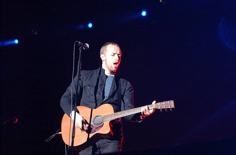 Chris Martin singing with an acoustic guitar under blue stage lights