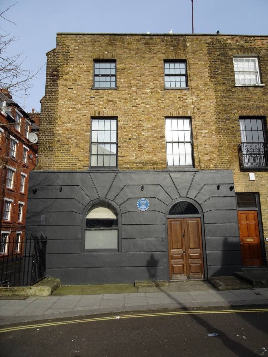 A three-storey yellow-brick building with a grey ground floor and a blue plaque