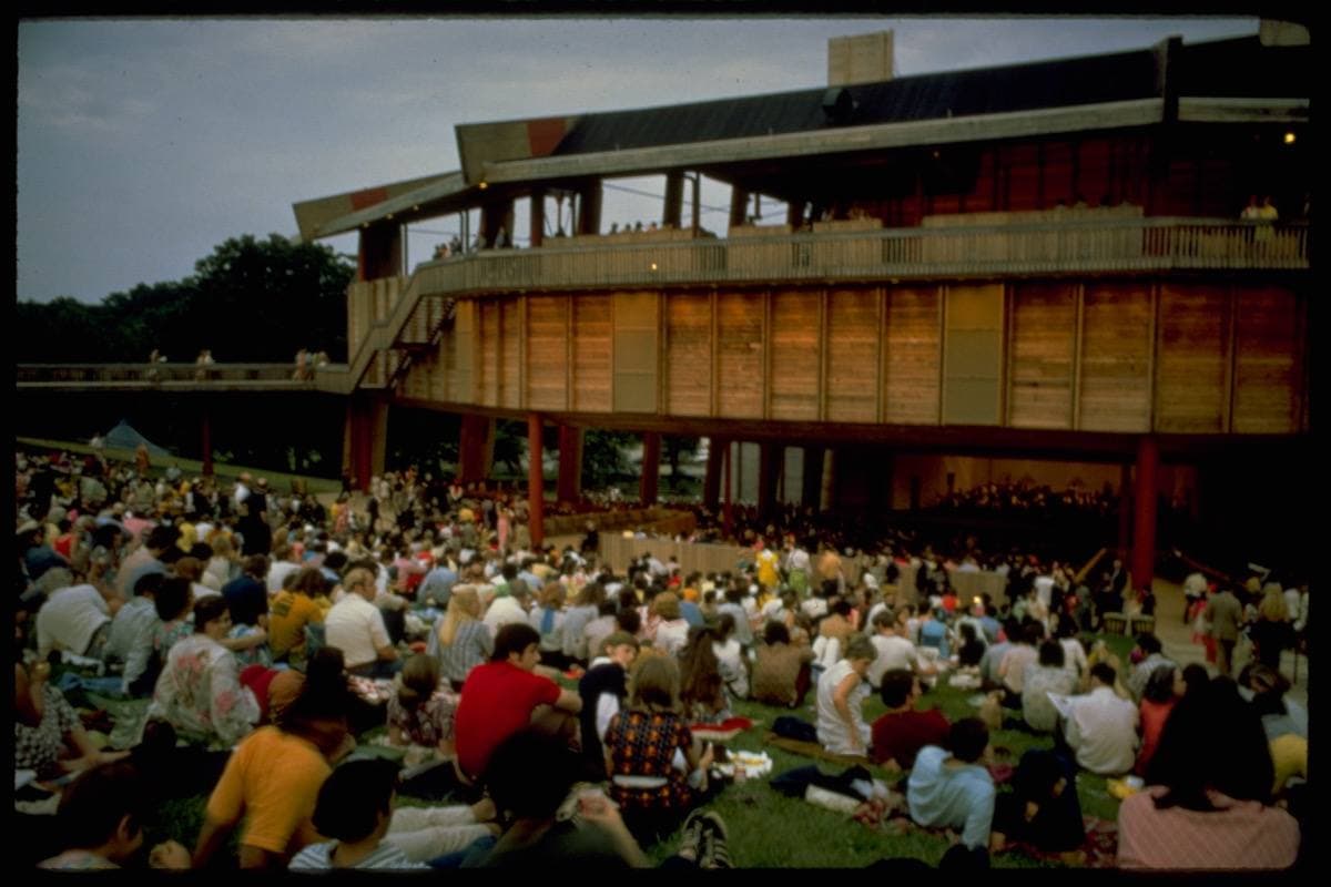 An open-sided timber concert pavilion with an audience on the lawn