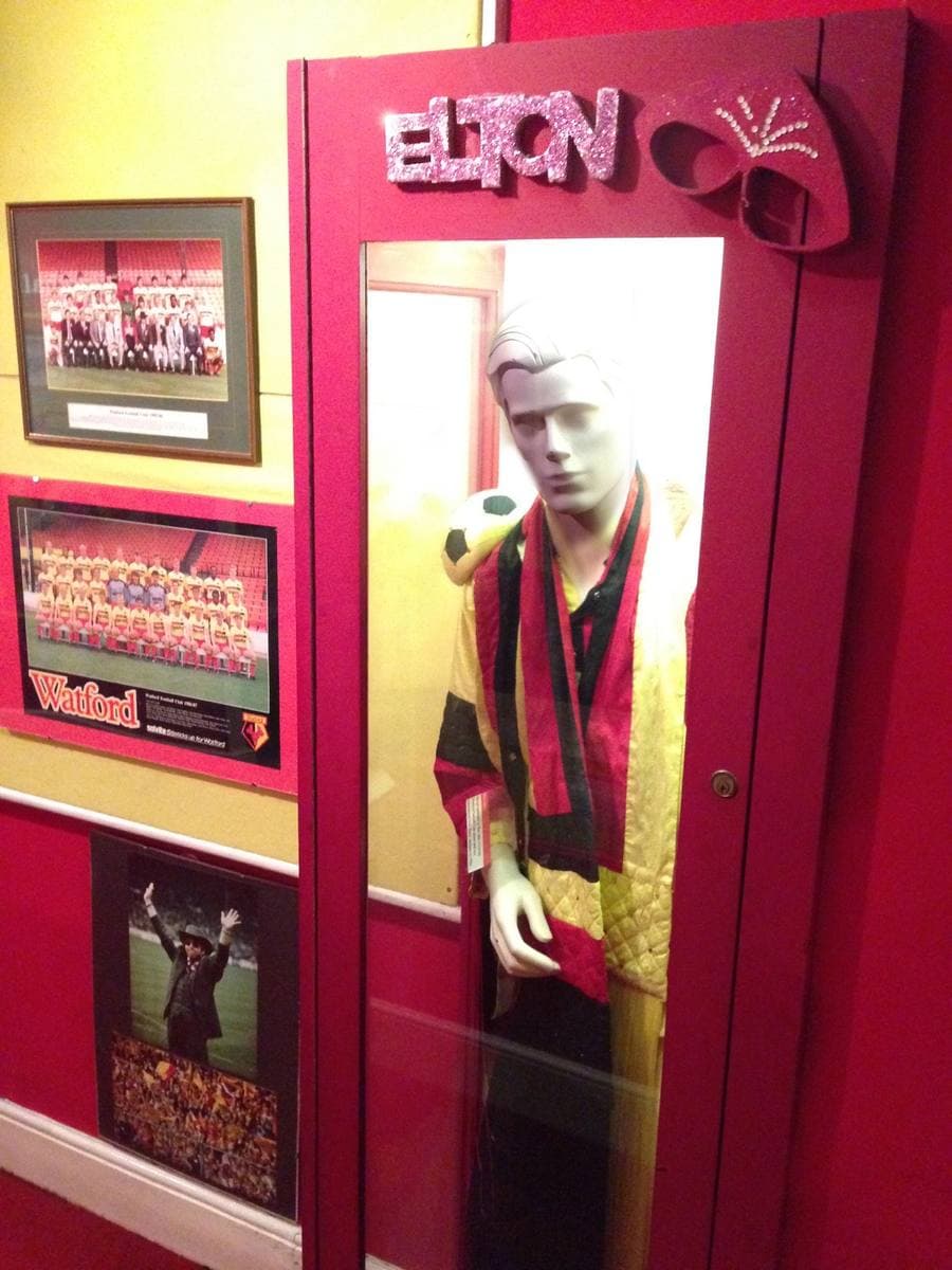 A mannequin in a quilted yellow, red and black costume and scarf inside a red display case topped with sparkly glasses and the word Elton
