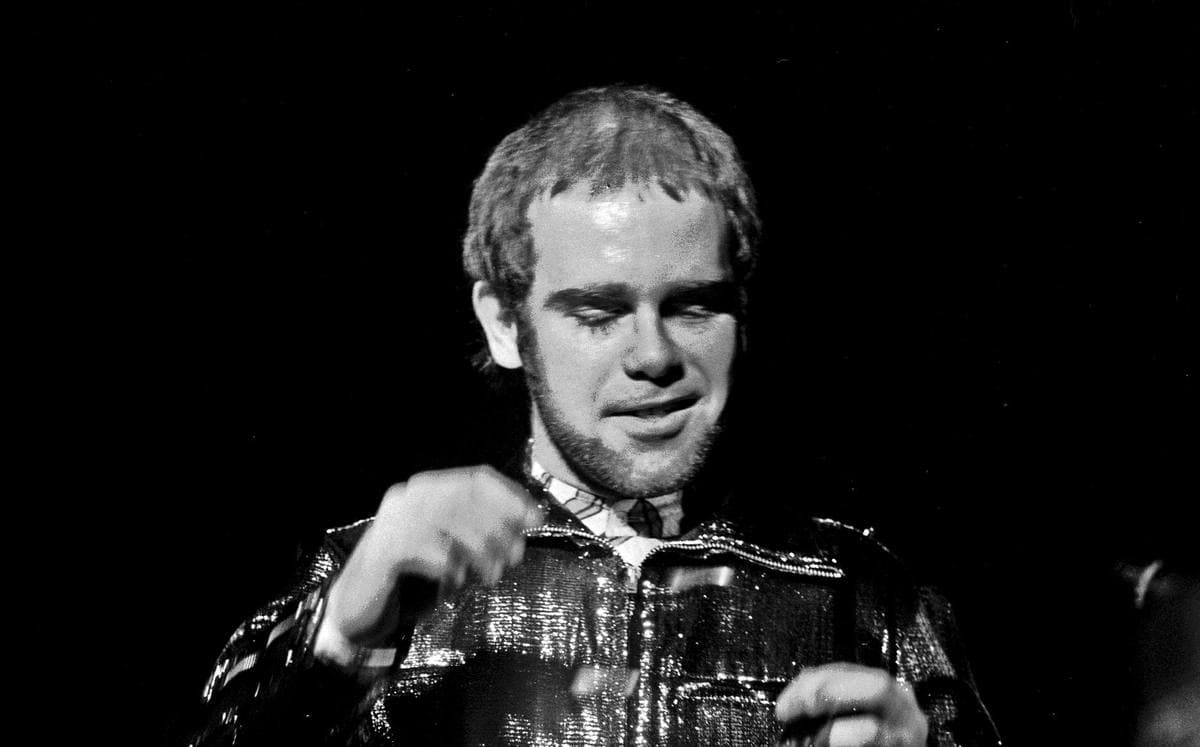 Elton John on stage in a glittering jacket, smiling, against a black background